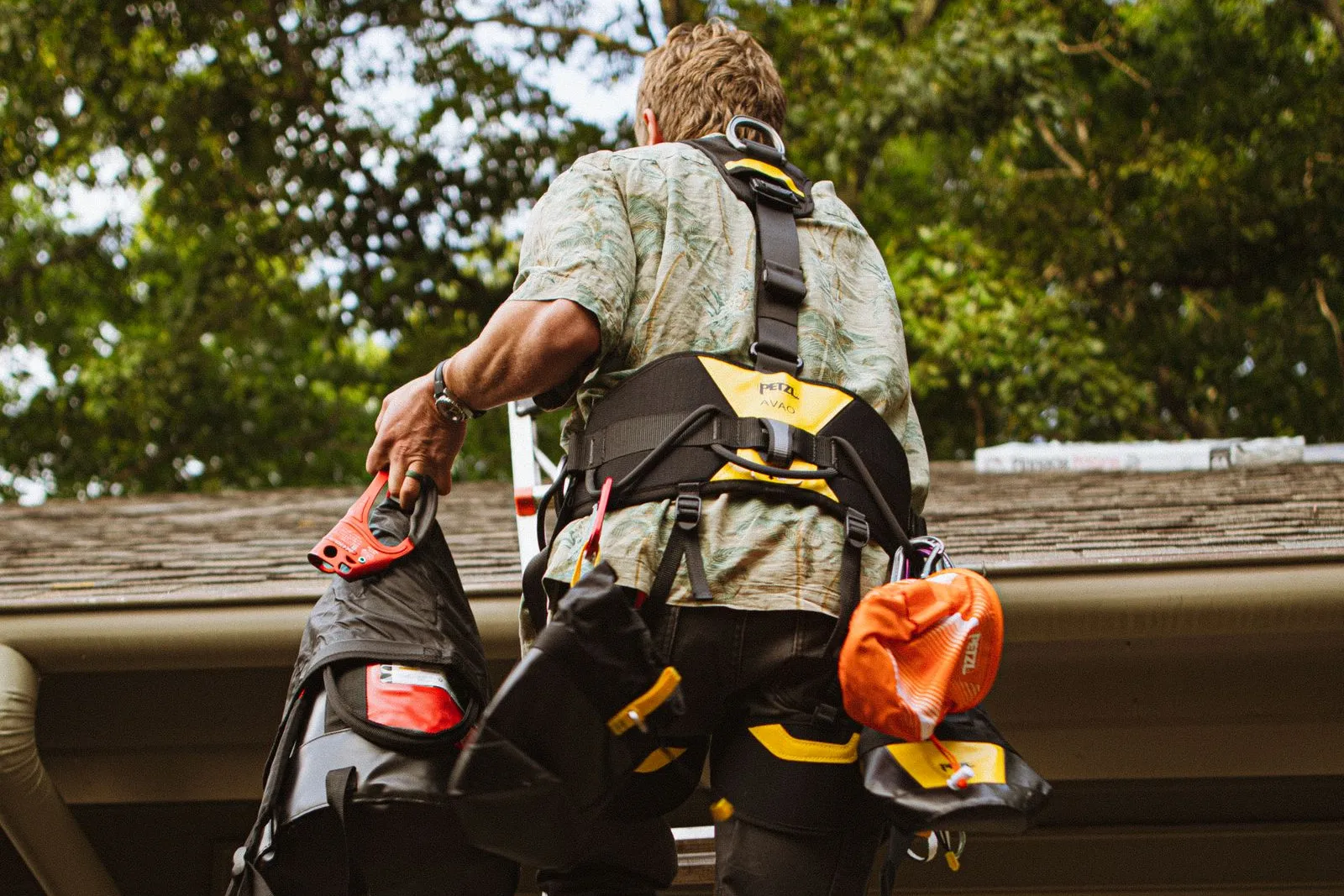 A worker in a safety harness climbs a ladder to a shingled roof